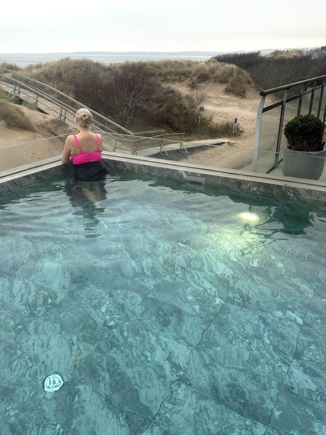 Elin syns bakifrån i utomhuspoolen. Hon har håret uppsatt i en knut och hennes baddräkt är rosa och svart. Hon tittar ut över havet. 